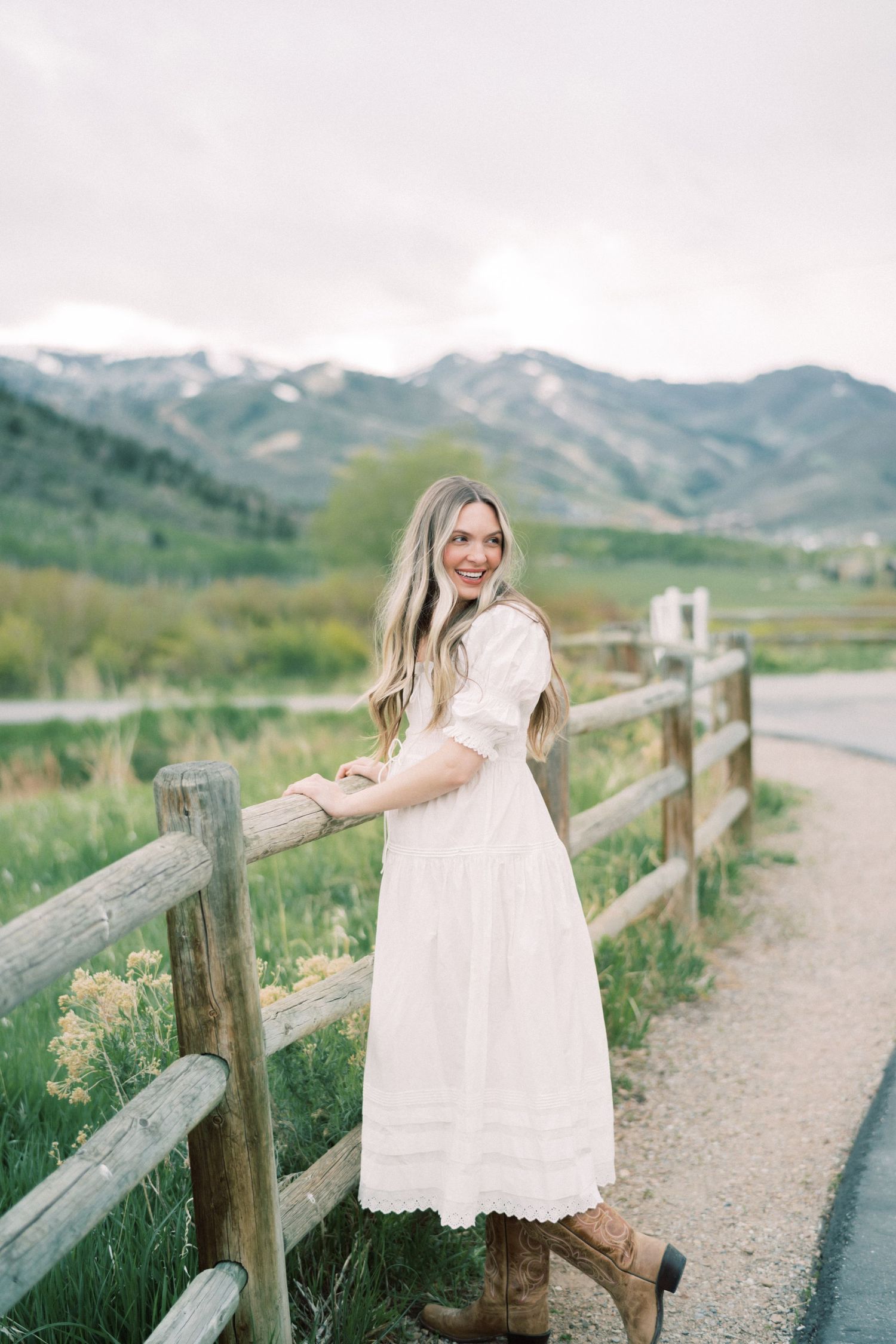 A woman in a white lace dress and boots leans on a wooden fence along a mountain path on a cloudy day.