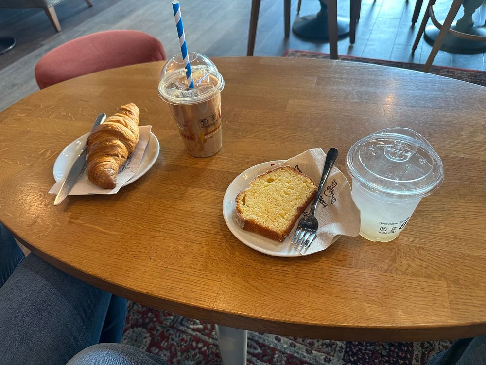 A wooden round cafe table with pastries, lemon loaf bread, and iced drinks served on white plates.