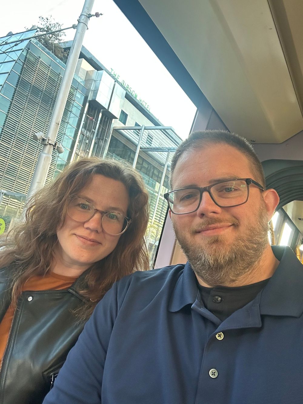 A selfie taken outdoors near a modern glass building showing two people wearing glasses in casual attire.