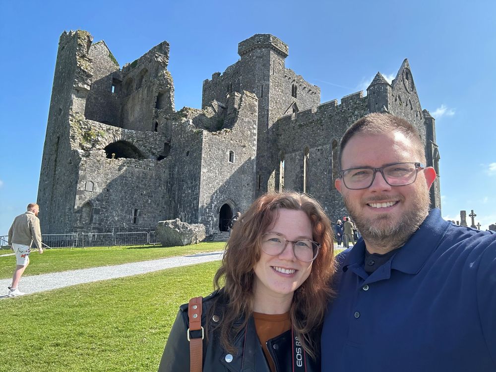 A stone castle ruin with tall towers stands in the background while visitors take a photo on a grassy lawn on a sunny day.