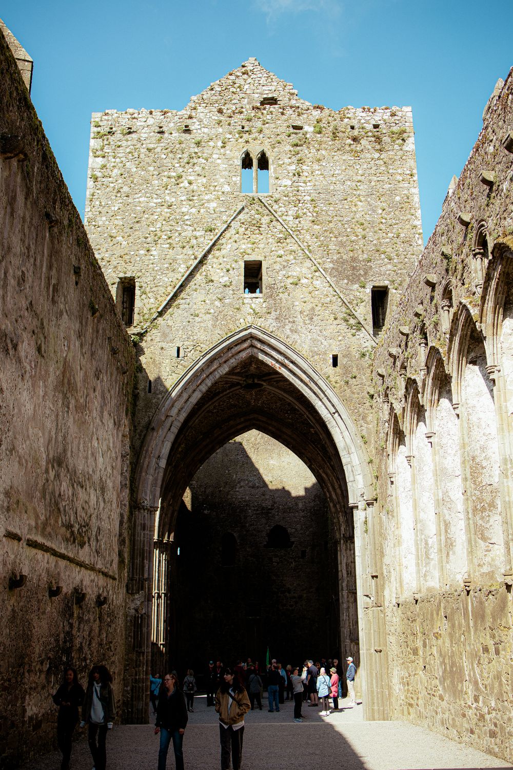 Medieval stone archway entrance and walls of a historic cathedral with pointed Gothic architecture and visitors walking beneath.