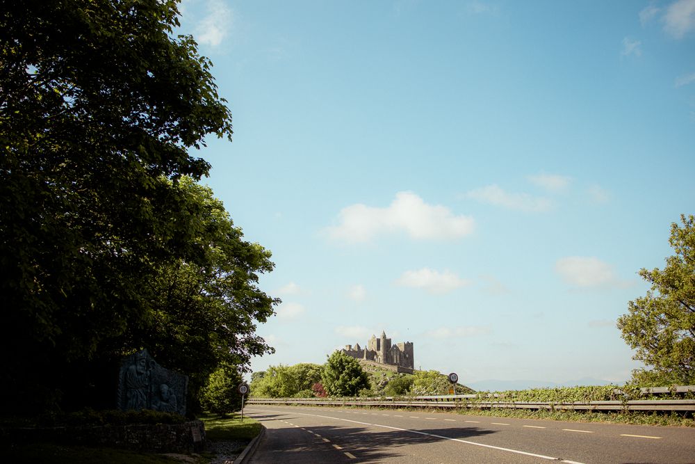 A winding road leads to a distant medieval castle silhouetted against a bright blue sky framed by green trees.