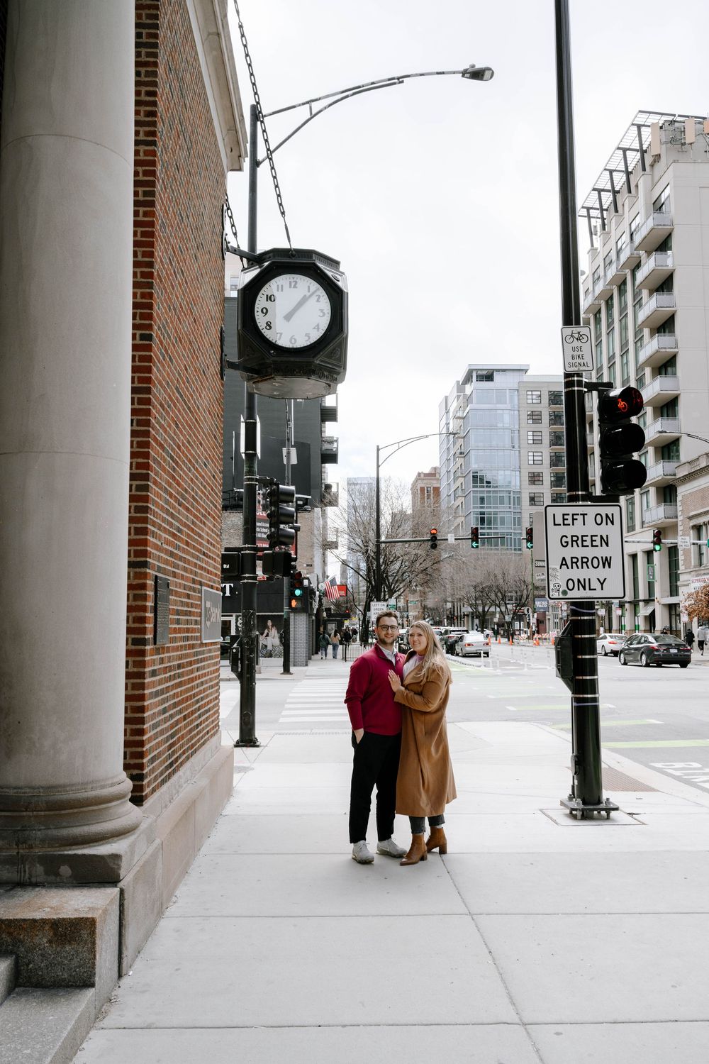 A couple embracing on a city street corner beneath a vintage clock and street sign on a winter afternoon.