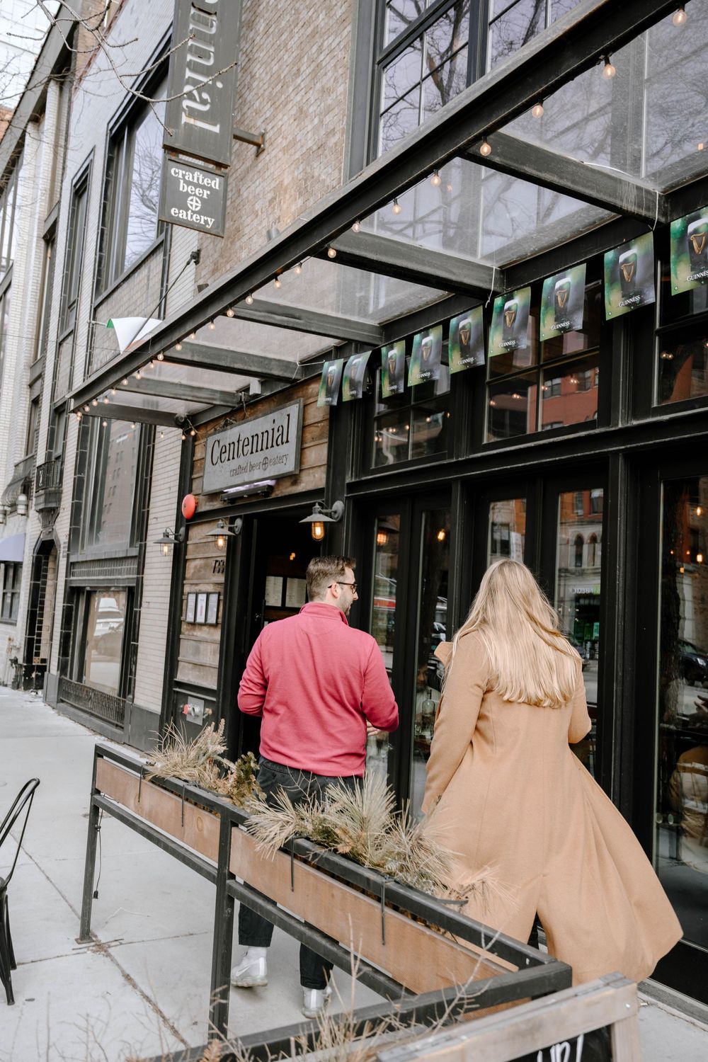 A couple walks by the exterior of Centennial craft beer eatery with a glass awning and rustic brick facade.