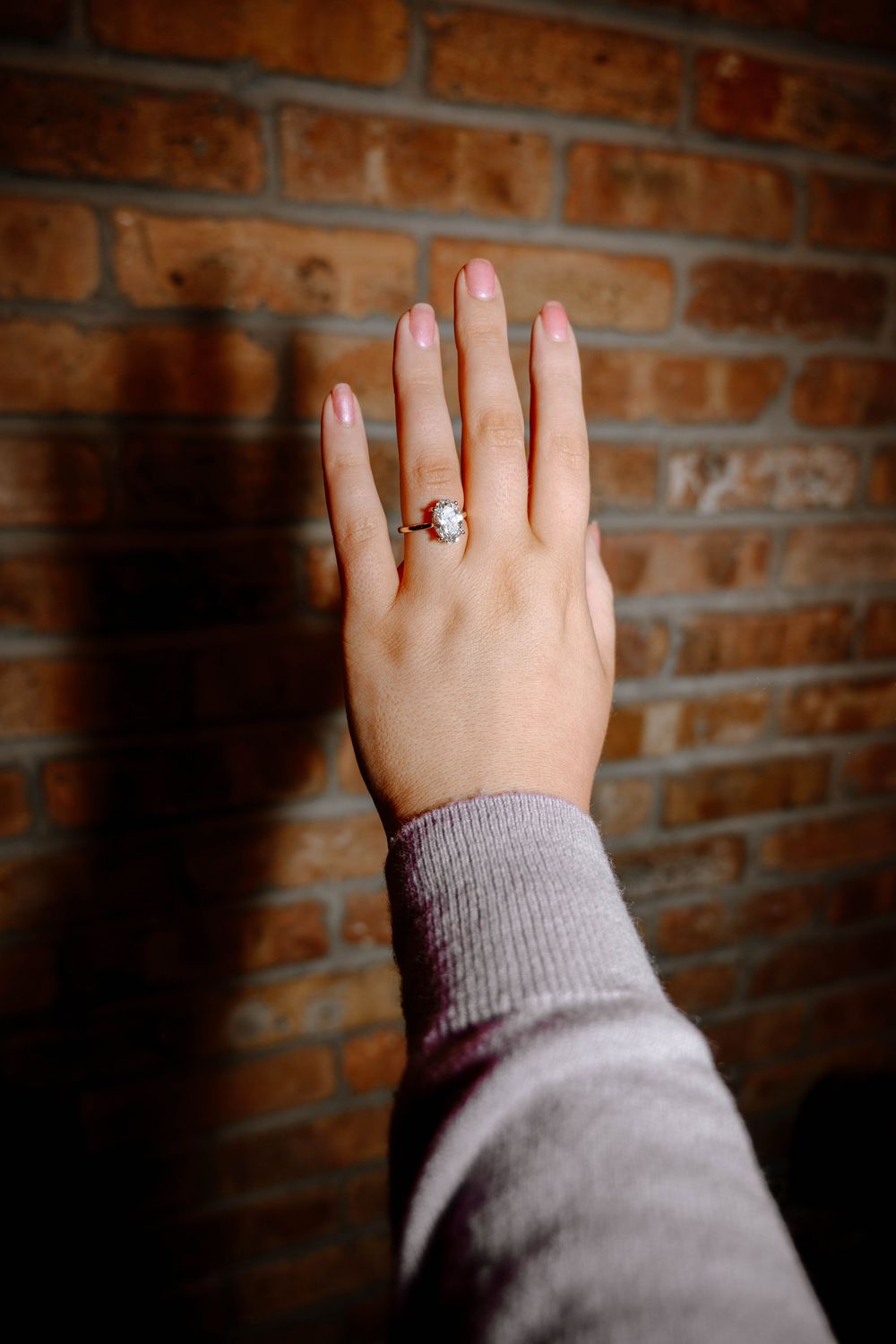 A hand wearing a silver ring with a stone setting against a rustic brick wall background.