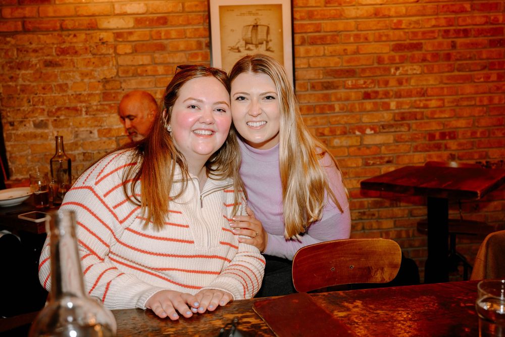 Two women smiling together at a restaurant with exposed brick walls and warm lighting.