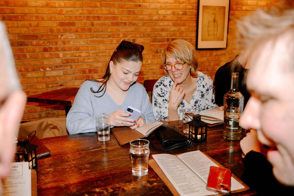 People gather around a wooden table at a bar or restaurant with brick walls, looking at phones and menus.