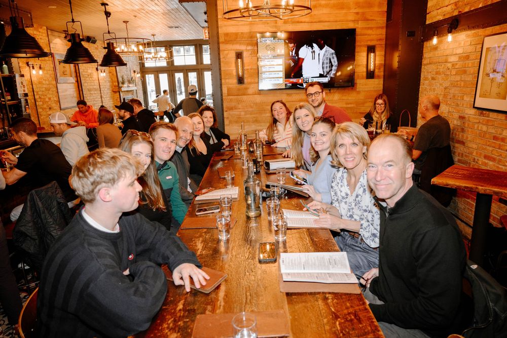 Large group dining together at a rustic wooden restaurant table with drinks and menus during a social gathering.