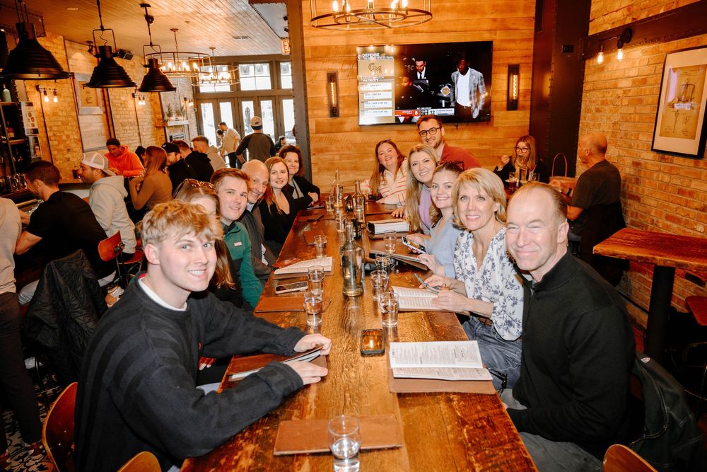 Group of diners seated at a long communal table inside a rustic restaurant with exposed brick walls and pendant lighting.