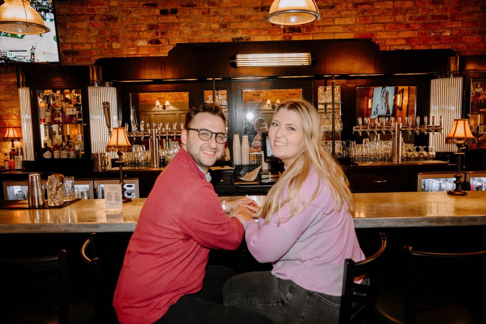 Two people sit together at a wooden bar counter inside a cozy pub with exposed brick walls and pendant lighting.