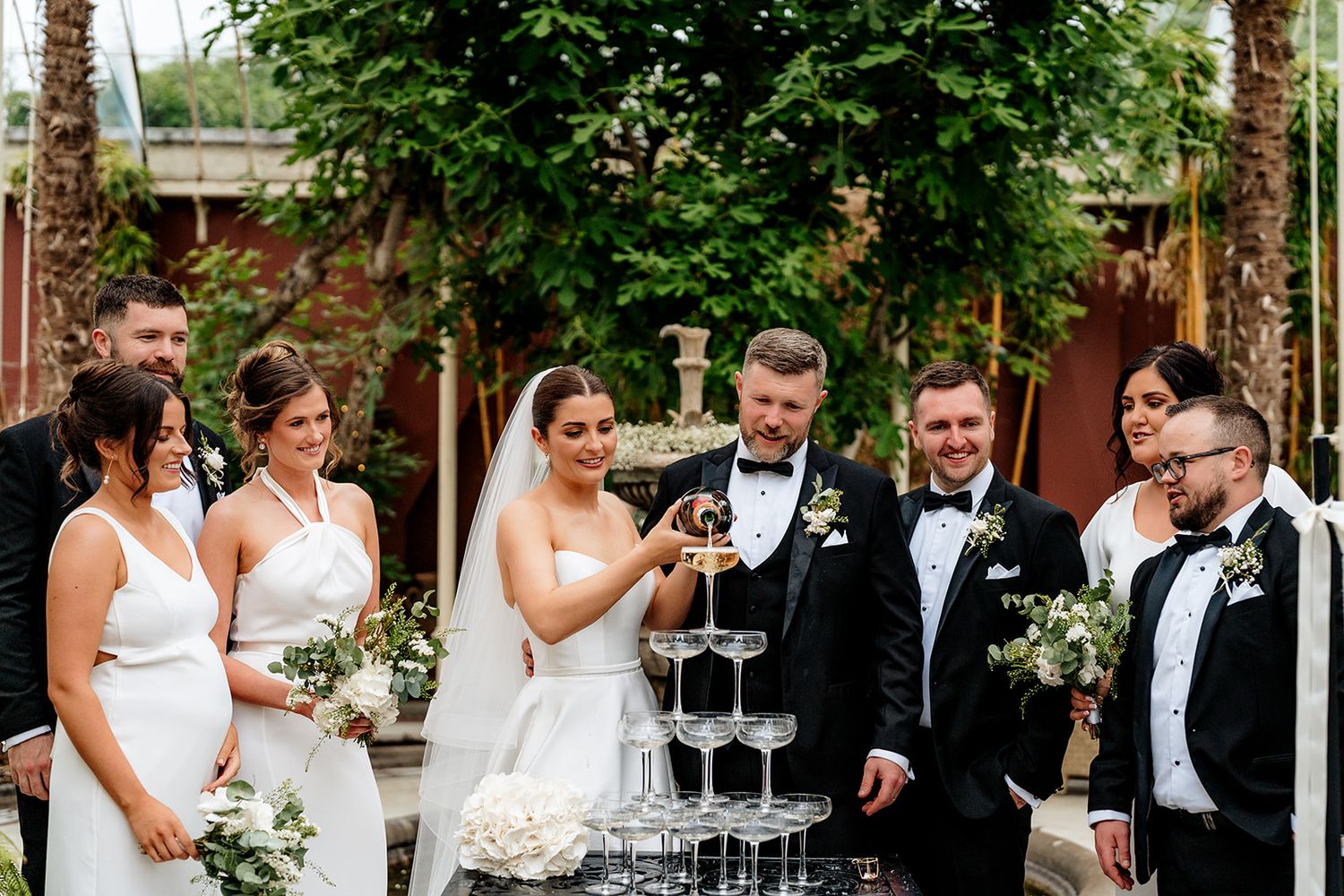 Wedding party gathers around champagne tower at outdoor reception venue with green trees in background.