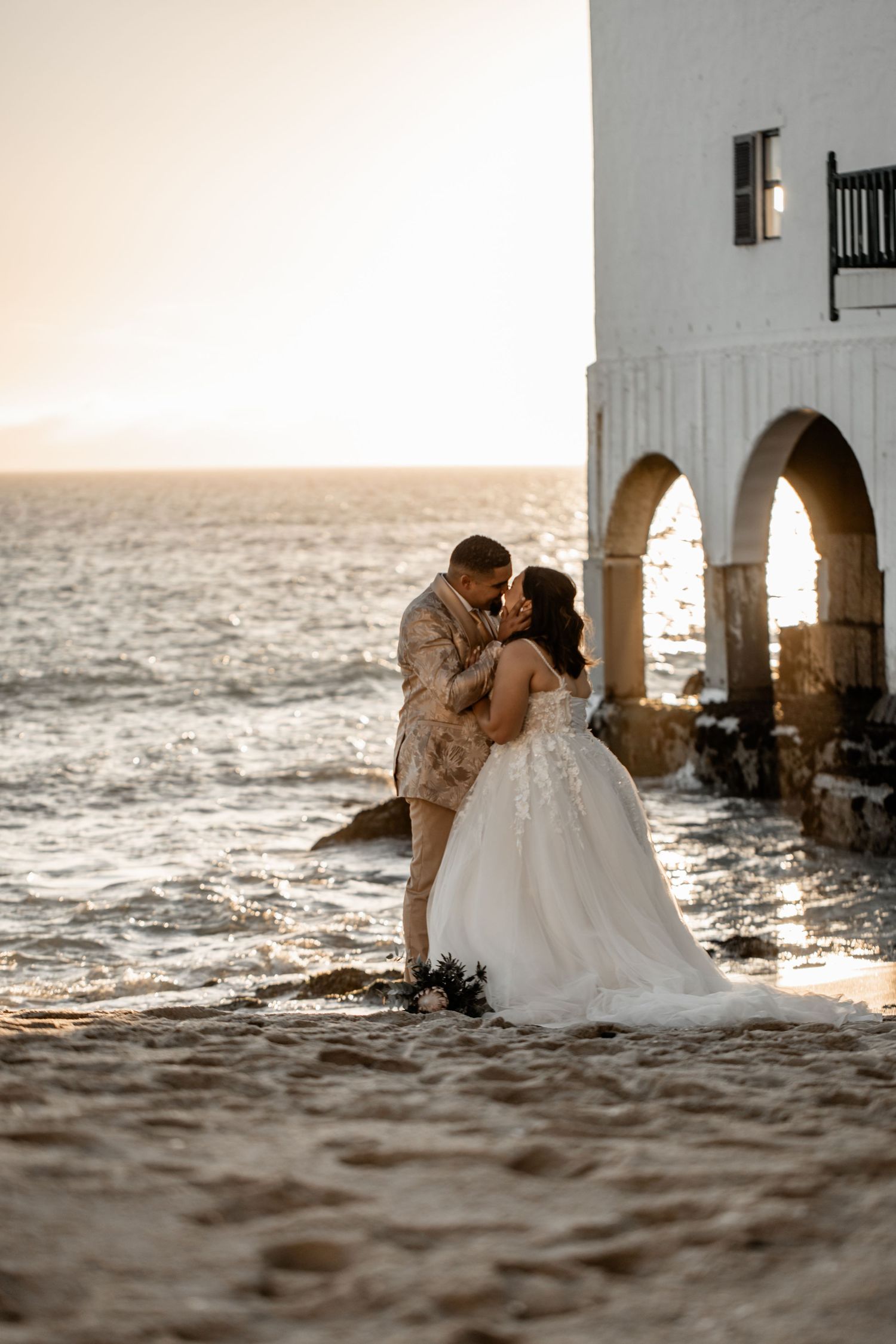 A romantic seaside wedding photo at sunset with a couple embracing beneath white Mediterranean arches.