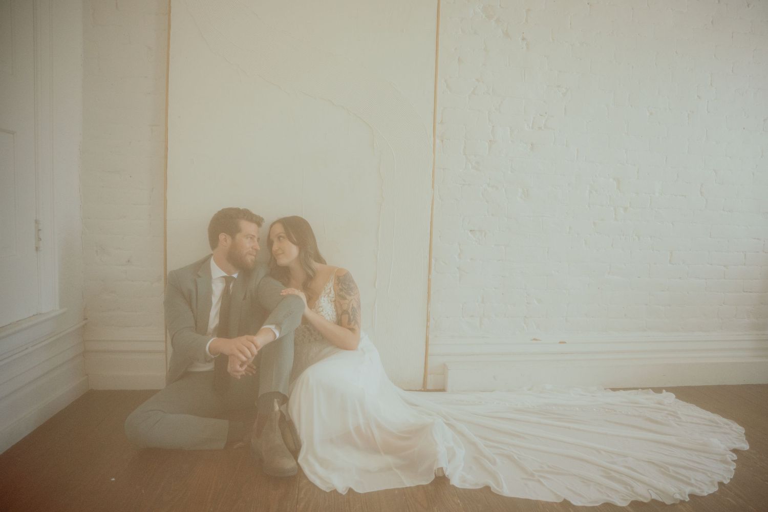 A bride and groom share an intimate moment while sitting against a white wall in dreamy lighting.