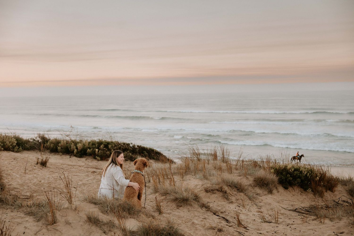 Couple sits together on sandy beach dunes overlooking misty ocean waves during golden sunset hour.