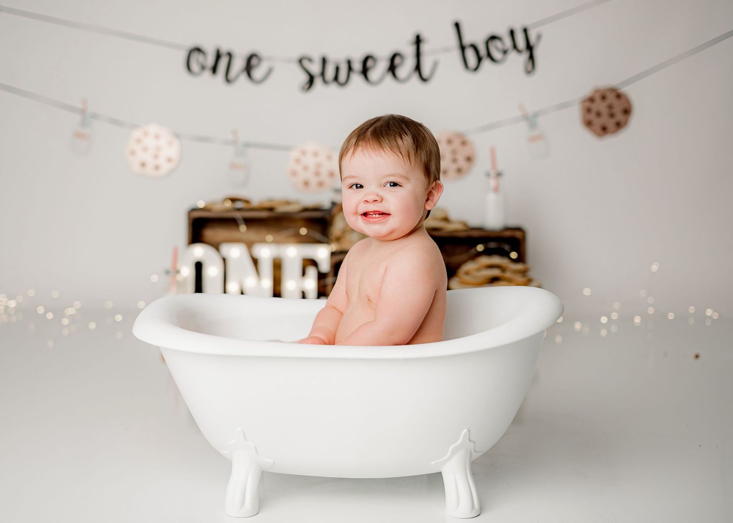 A smiling baby sits in a white vintage bathtub with decorative lights and copper snowflakes in the background.