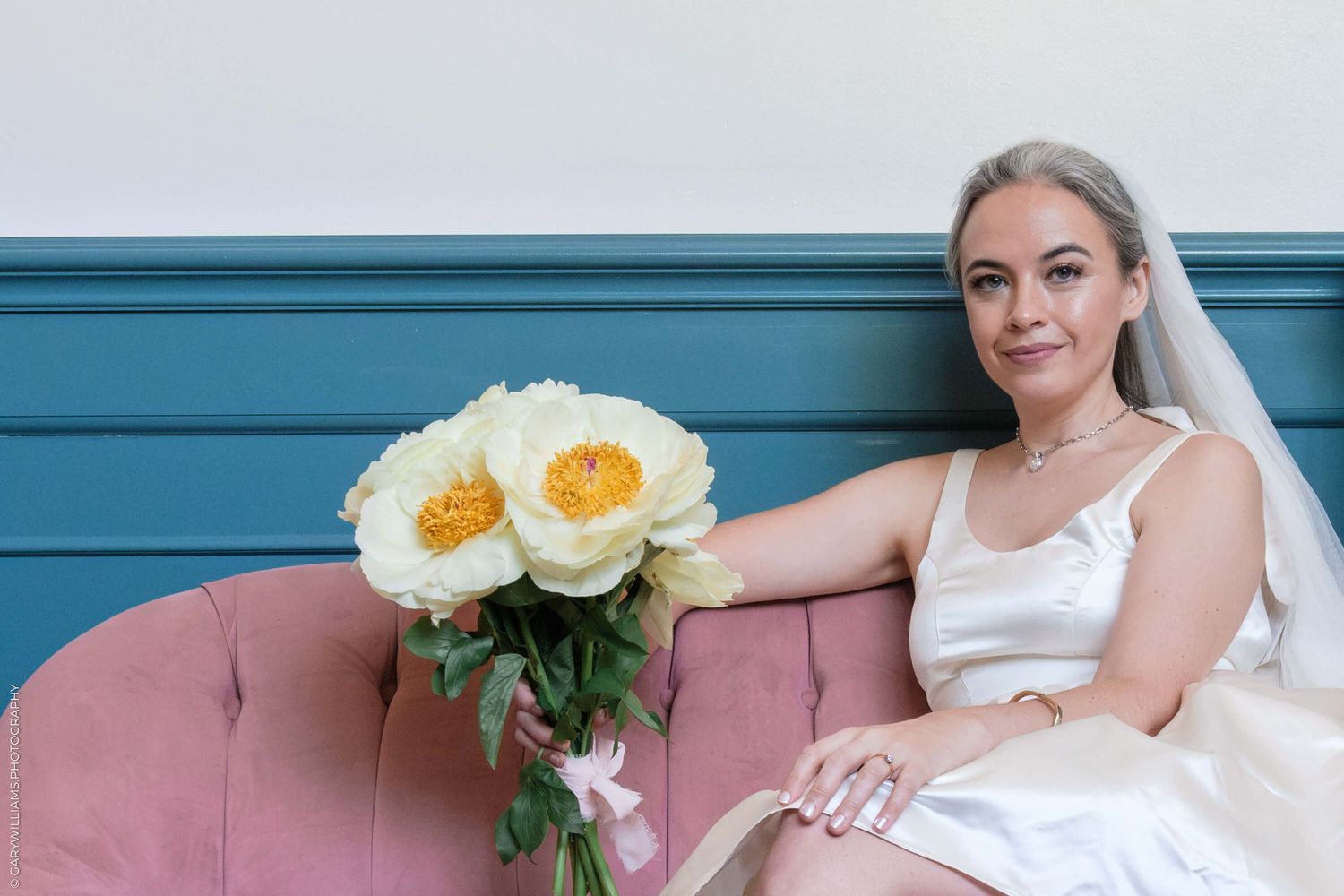 A bride in white dress sits on a pink sofa holding white and yellow peony bouquet against teal-colored wall.