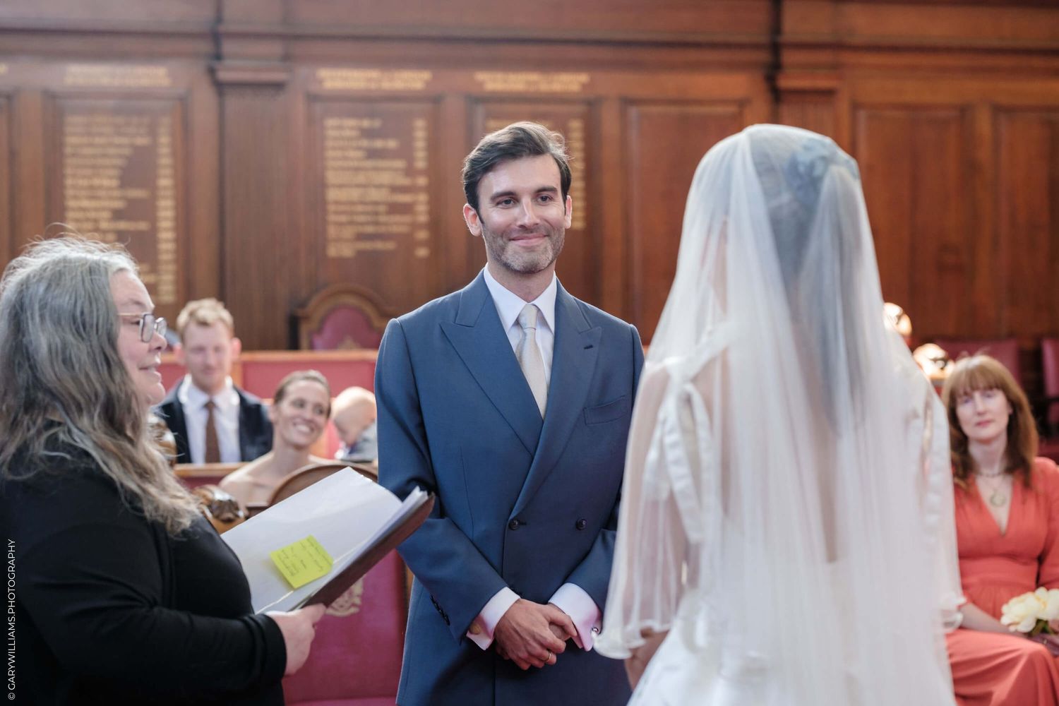 Wedding ceremony in a wood-paneled room shows groom in blue suit with officiant and bride during the service.