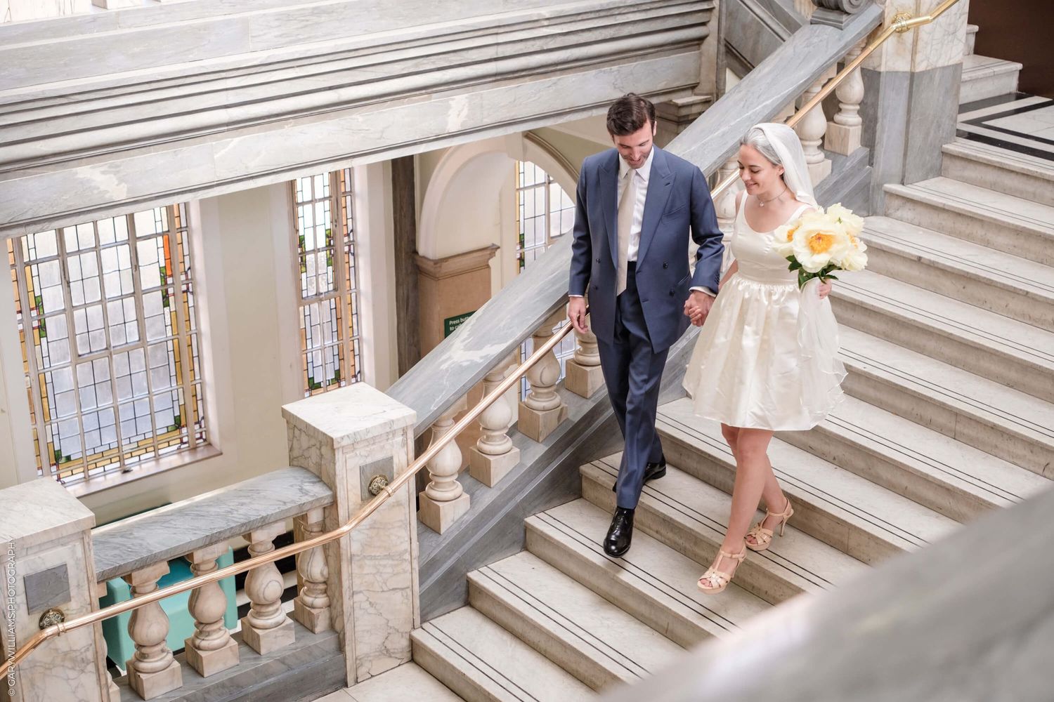 Couple walks down elegant marble staircase at historic courthouse venue in formal attire holding yellow bouquet.
