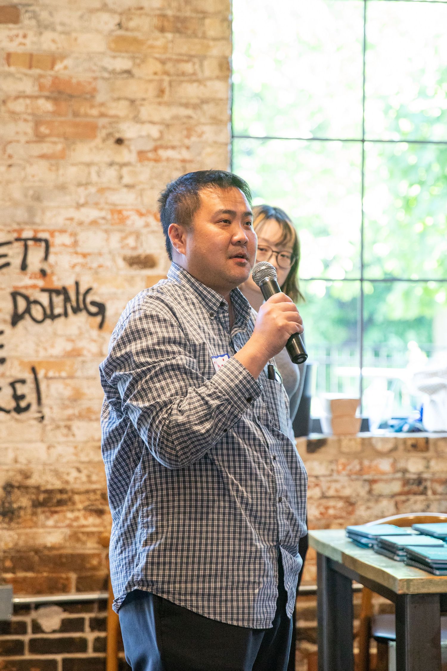 Speaker holds microphone while presenting in front of exposed brick wall in casual business setting.