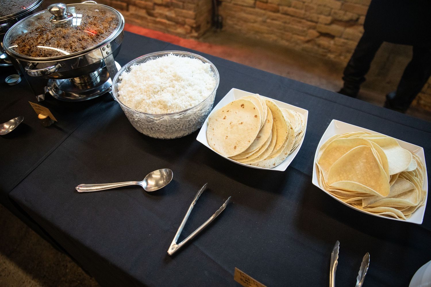 A buffet table with a rice bowl, serving utensils, and two plates of tortillas on a black tablecloth.