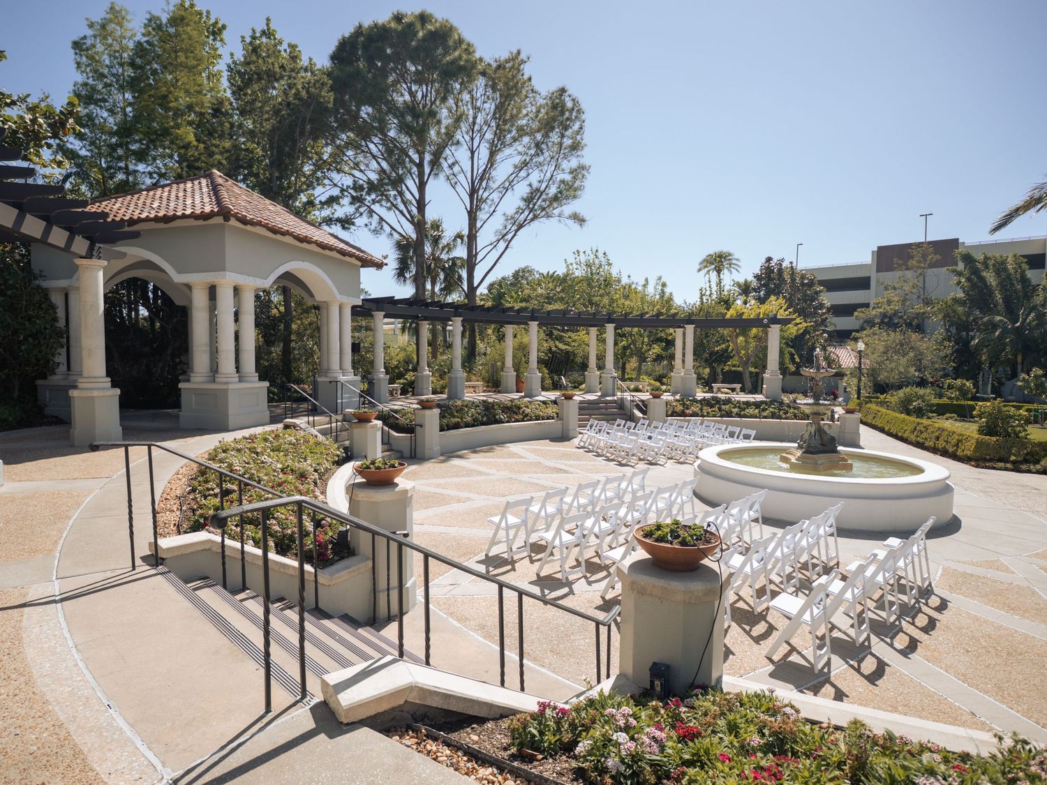 Elegant outdoor wedding venue with white chairs arranged in rows facing a gazebo amid manicured gardens and palm trees.