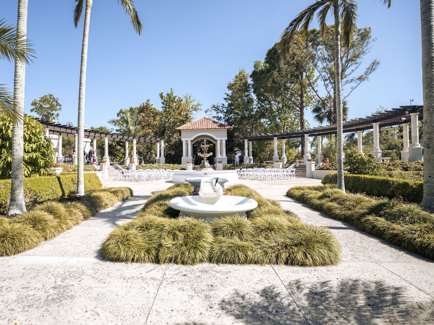 Elegant wedding venue courtyard with white fountain centerpiece surrounded by manicured gardens and tall palm trees.