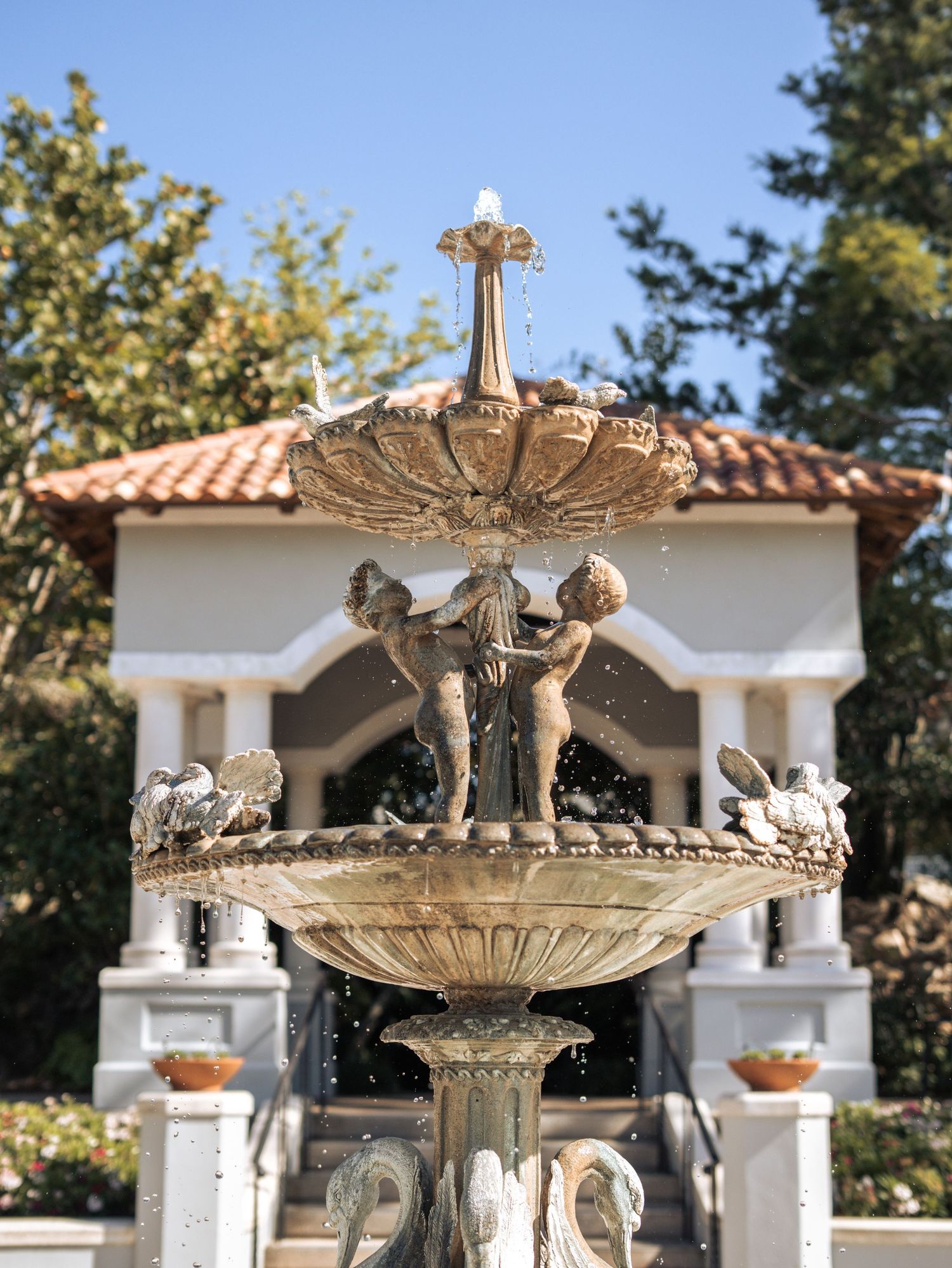 Ornate stone fountain with cherub sculptures and decorative crown top sits in front of a white Mediterranean-style building.