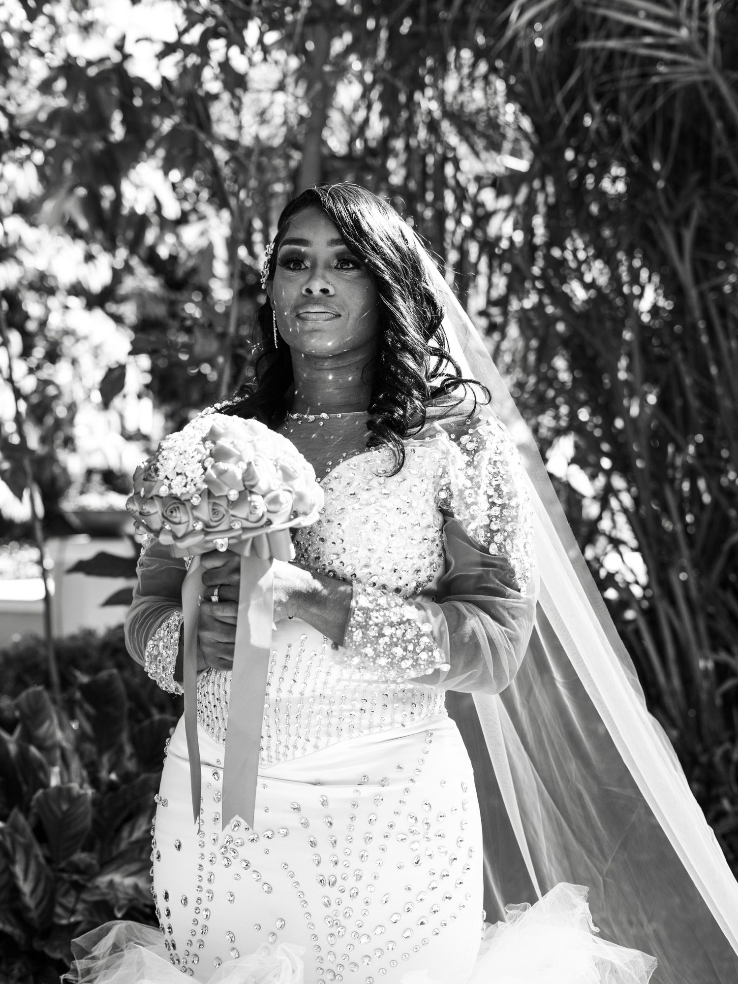 A stunning black and white bridal portrait showcasing an elegant lace wedding dress with a flowing veil in a garden setting.