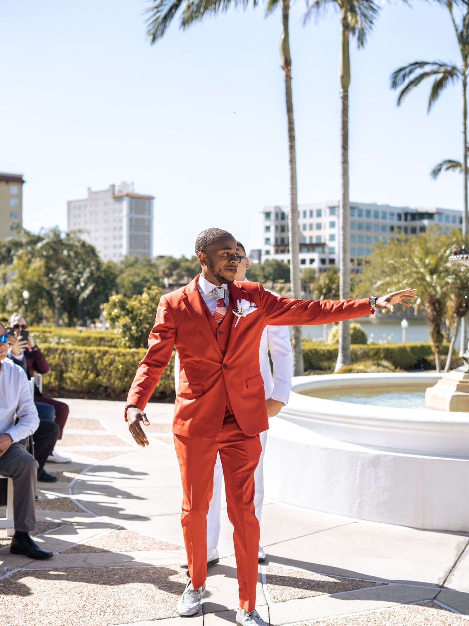 A person in a vibrant red suit strikes a stylish pose on a sunny terrace with palm trees and city buildings in the background.