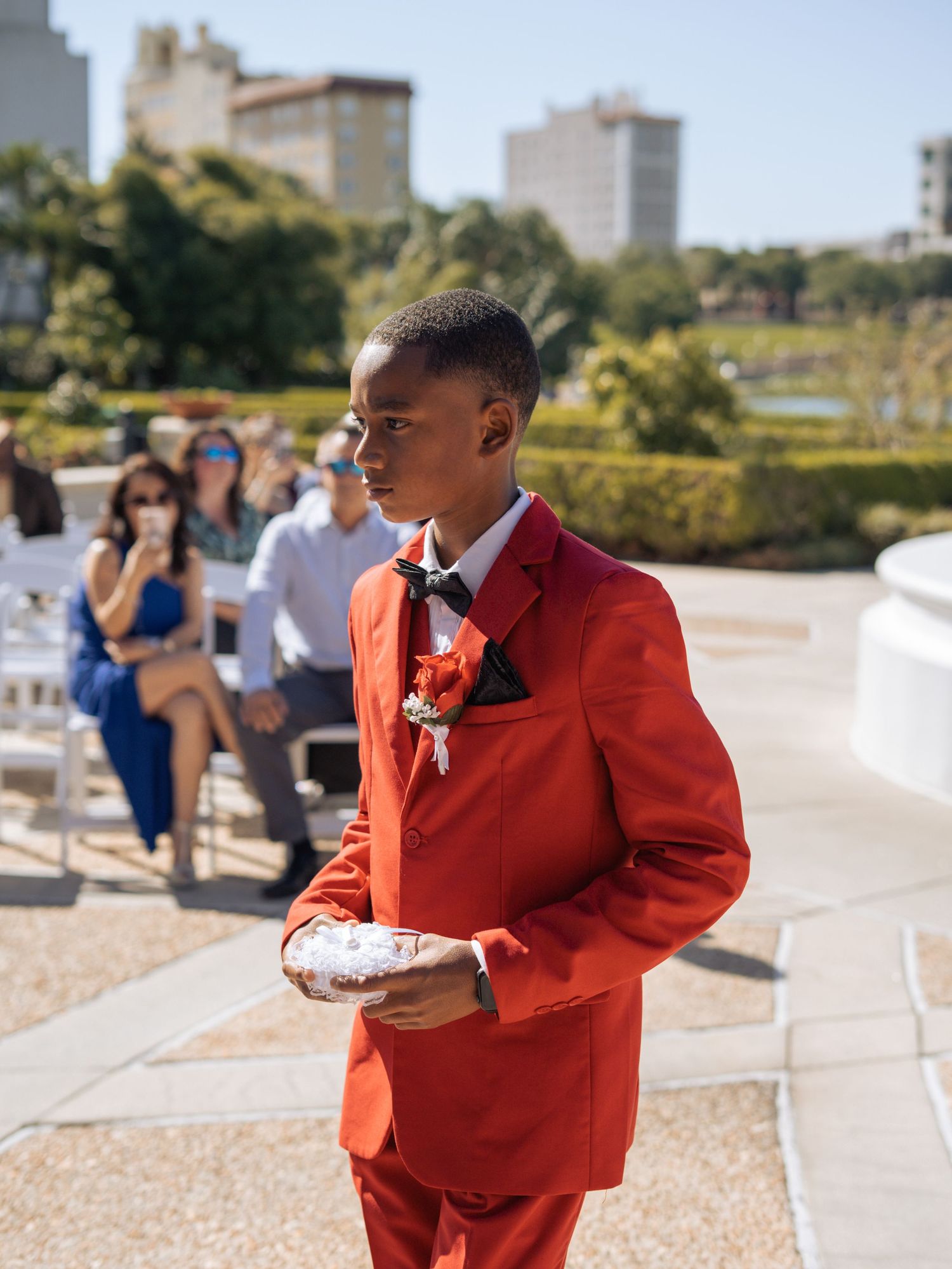 Groomsman in bright orange suit holding ring bearer pillow during outdoor ceremony.