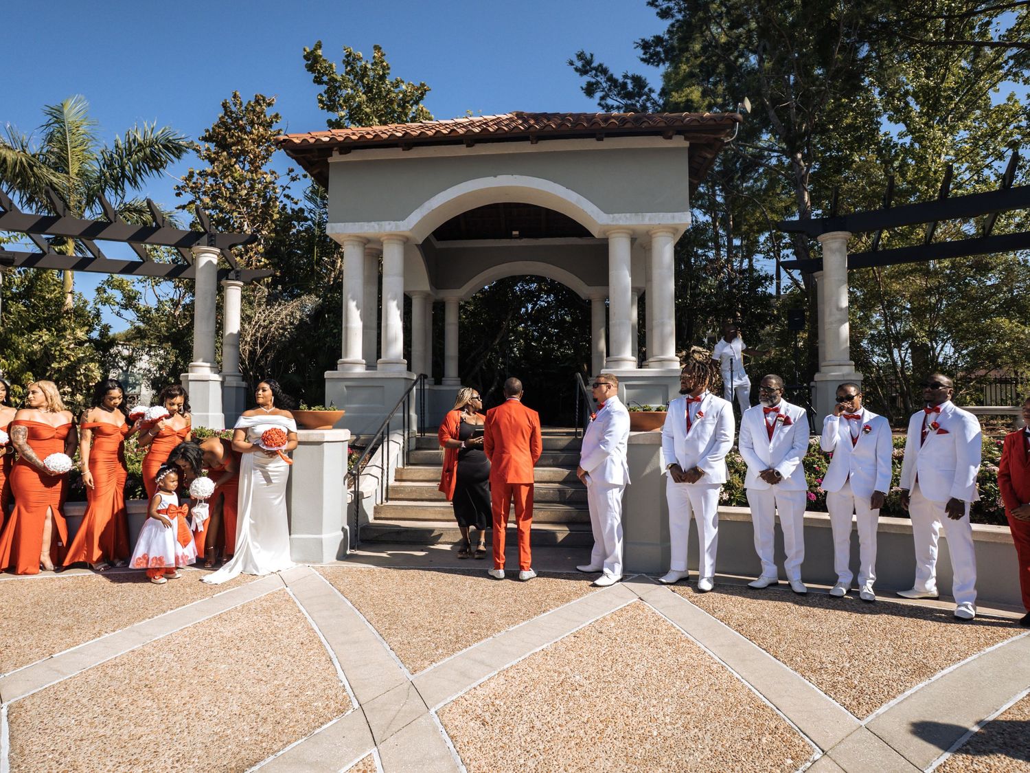 A wedding party lineup at an elegant gazebo with bridesmaids in orange dresses and groomsmen in white suits.
