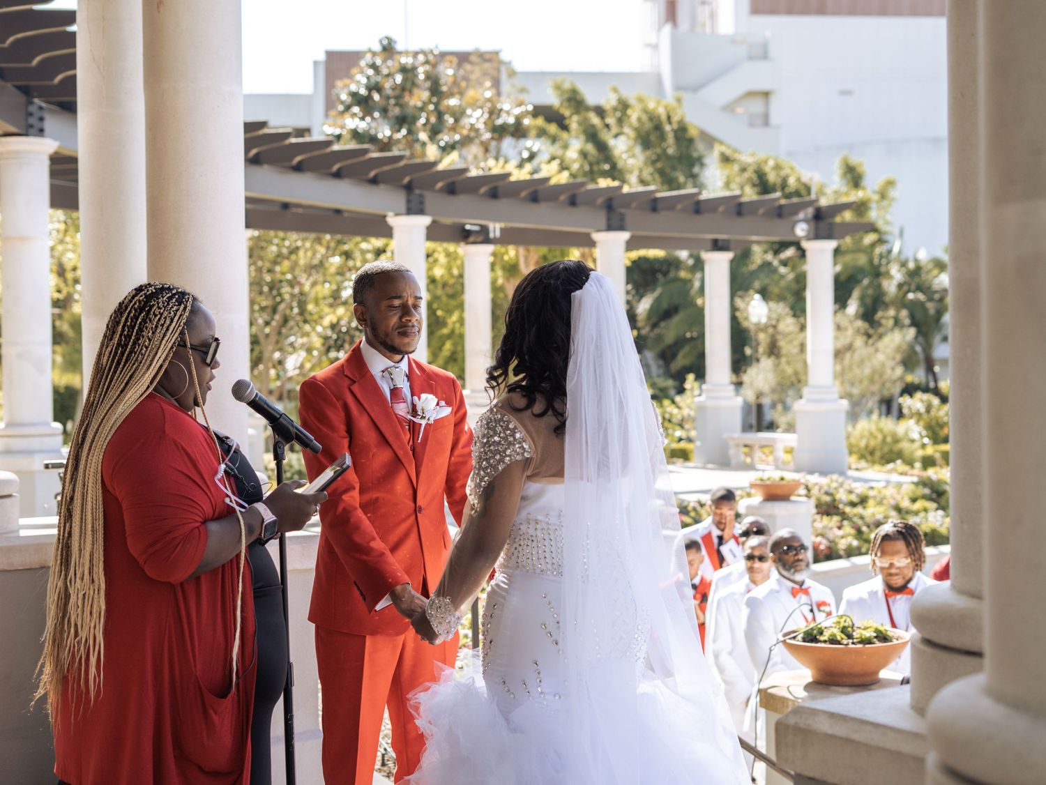 A wedding ceremony taking place under a column-lined pergola with floral decorations.