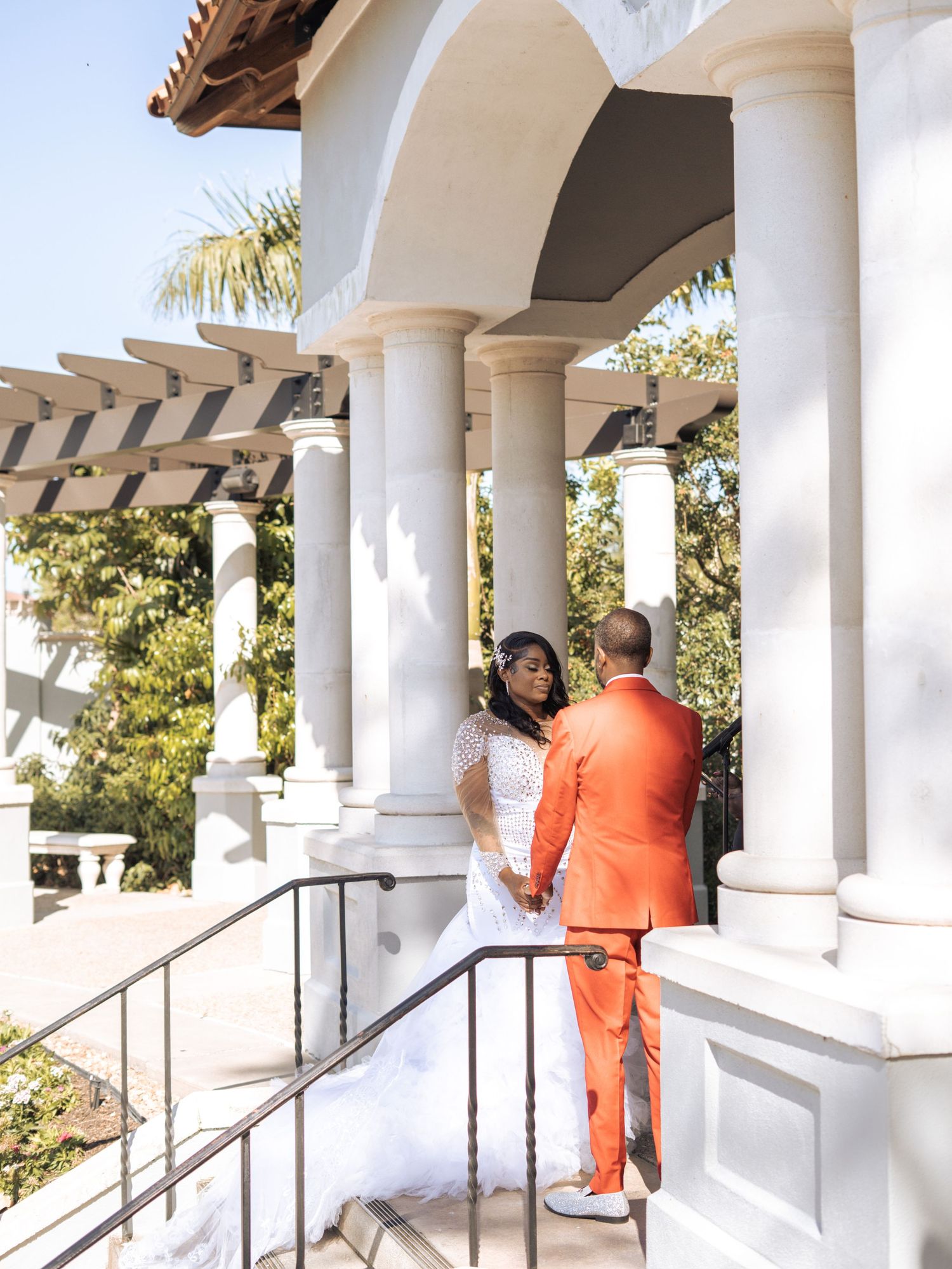 A wedding couple stands on steps under elegant white columns and arches at an outdoor venue.