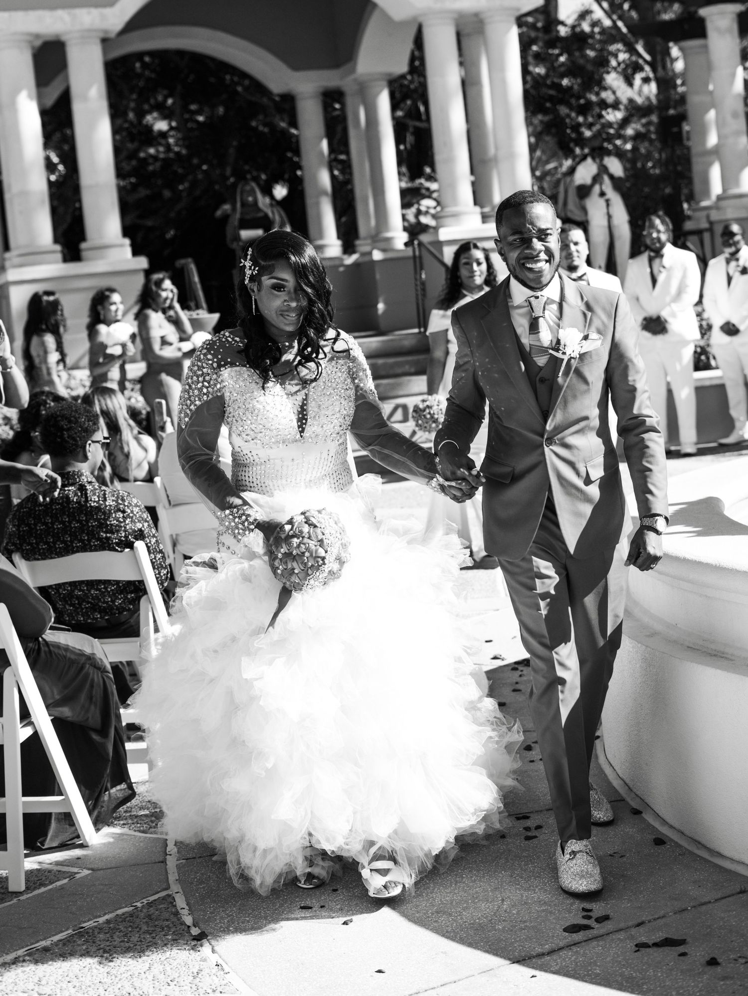 A joyful celebration moment at an outdoor wedding ceremony with white columns and guest seating.