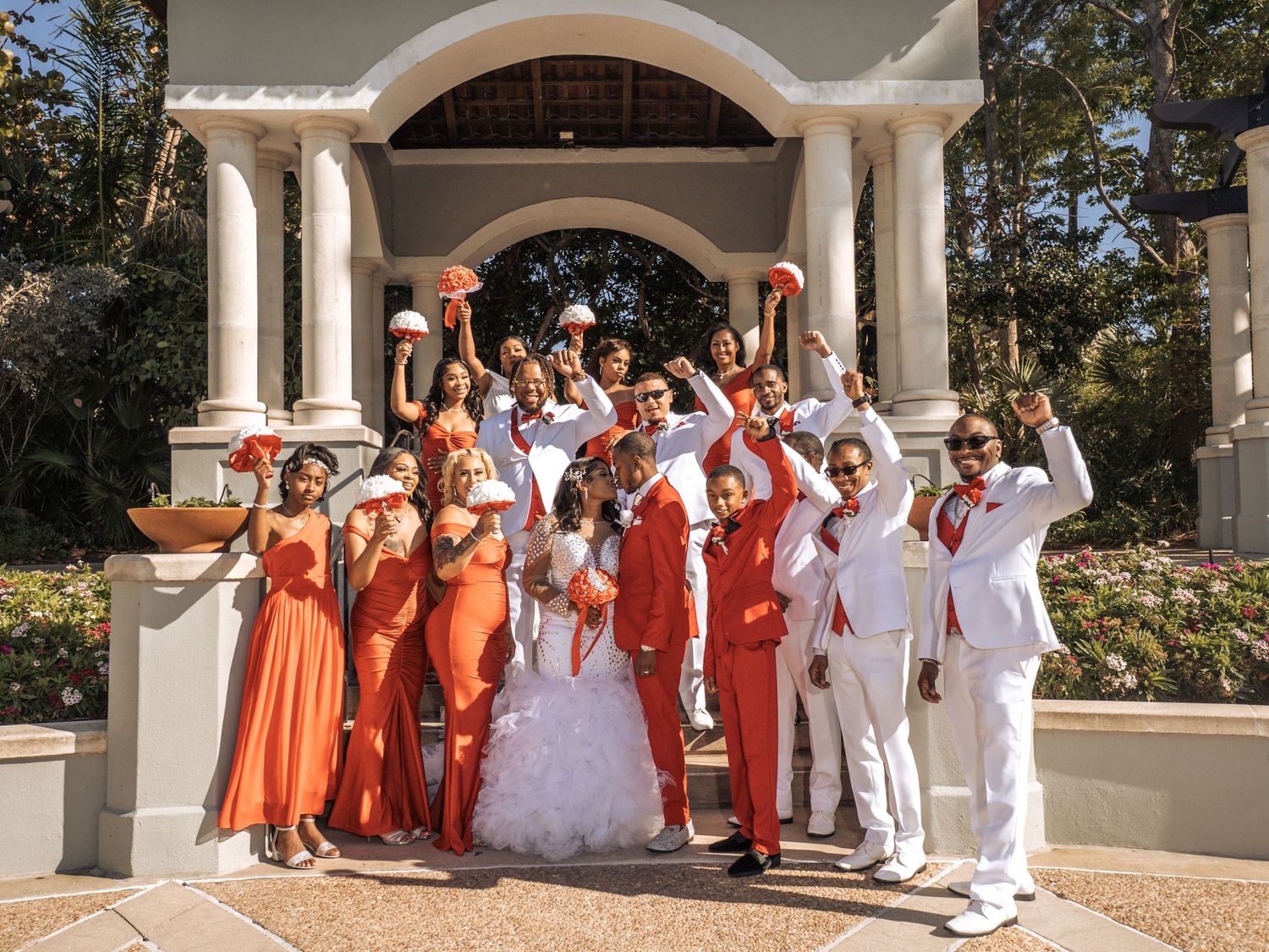 Wedding party in orange and white attire poses under an elegant arched gazebo on a sunny day.