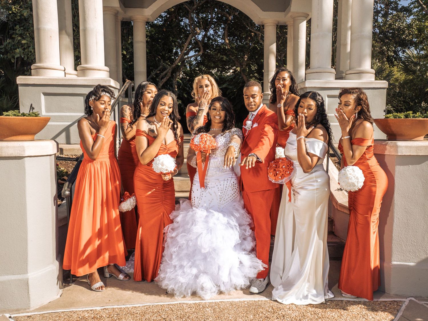 A wedding party wearing coral and orange bridesmaid dresses poses together with bouquets on an outdoor veranda.