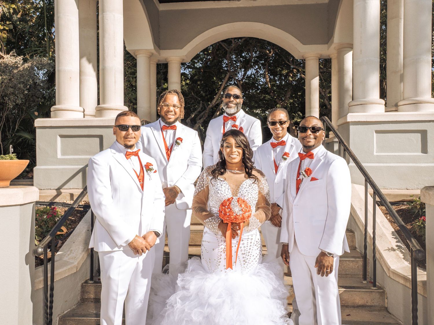 A wedding party wearing crisp white suits and a white gown pose on elegant stone steps with white columns outdoors.
