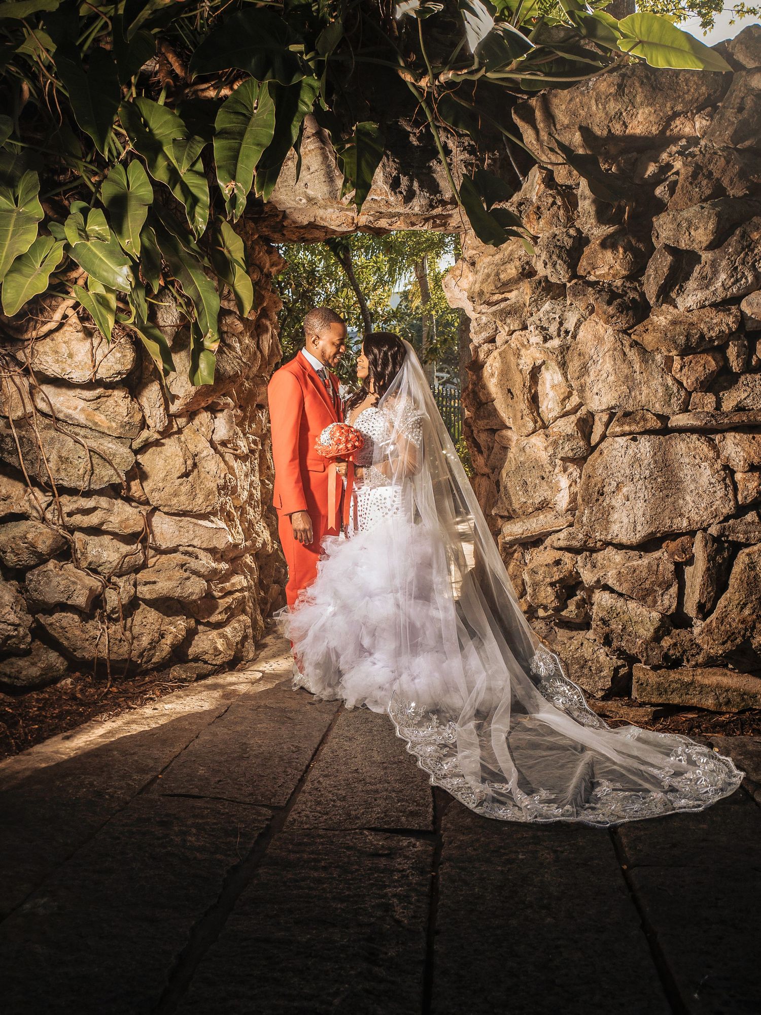A wedding couple in romantic evening light stands beneath a stone archway with a flowing white veil trailing behind.