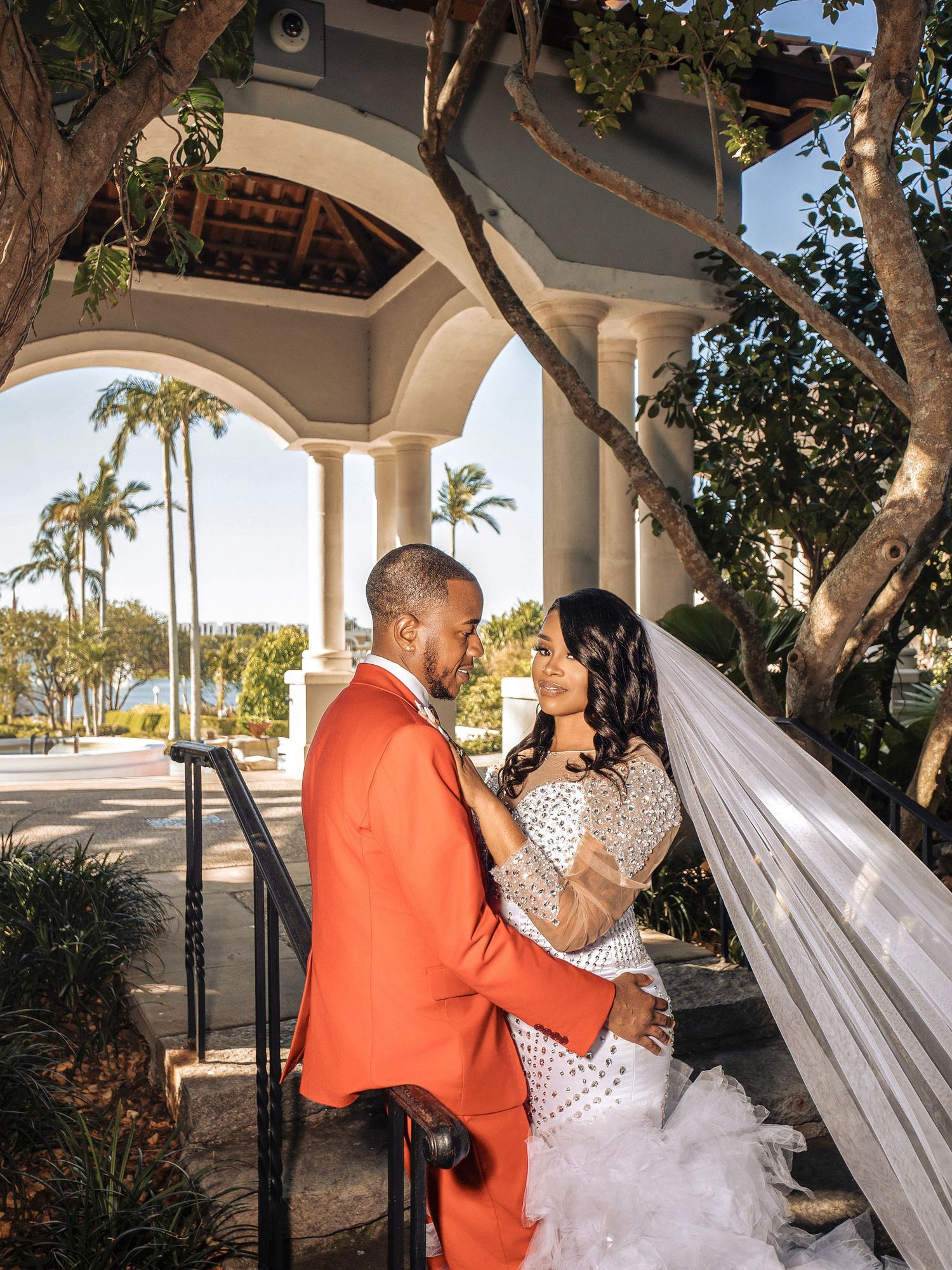 A elegant wedding couple embraces under an arched walkway with palm trees and sunset lighting in a tropical setting.