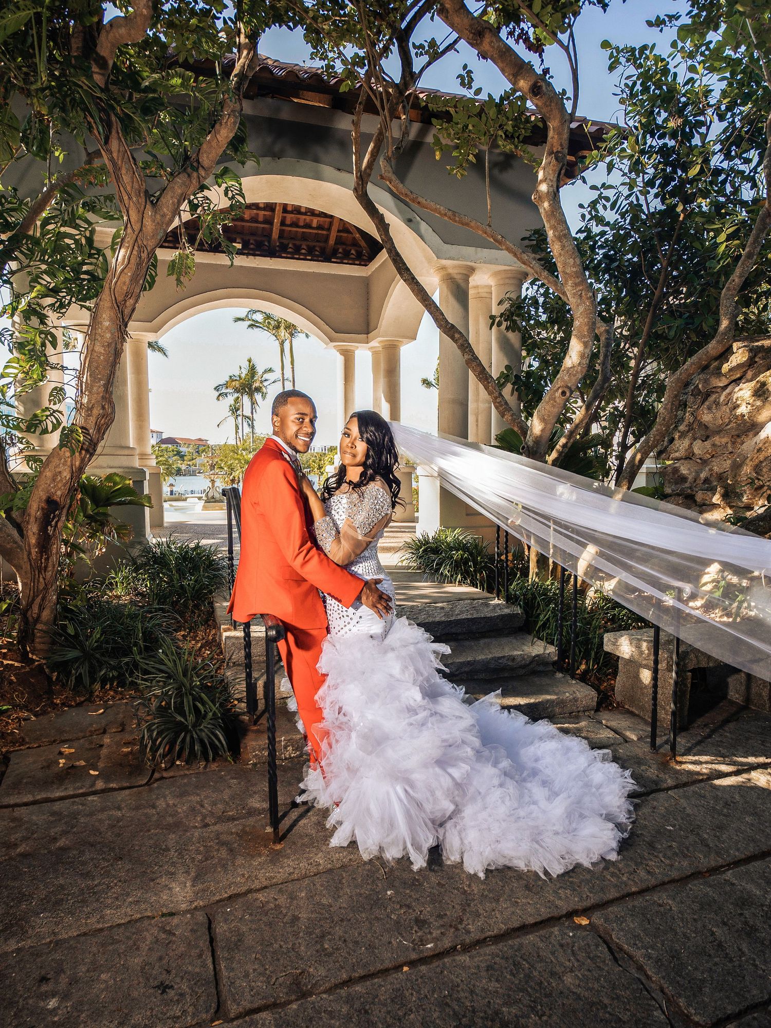 A couple poses under an elegant arched walkway with tropical palm trees in the background on their wedding day.