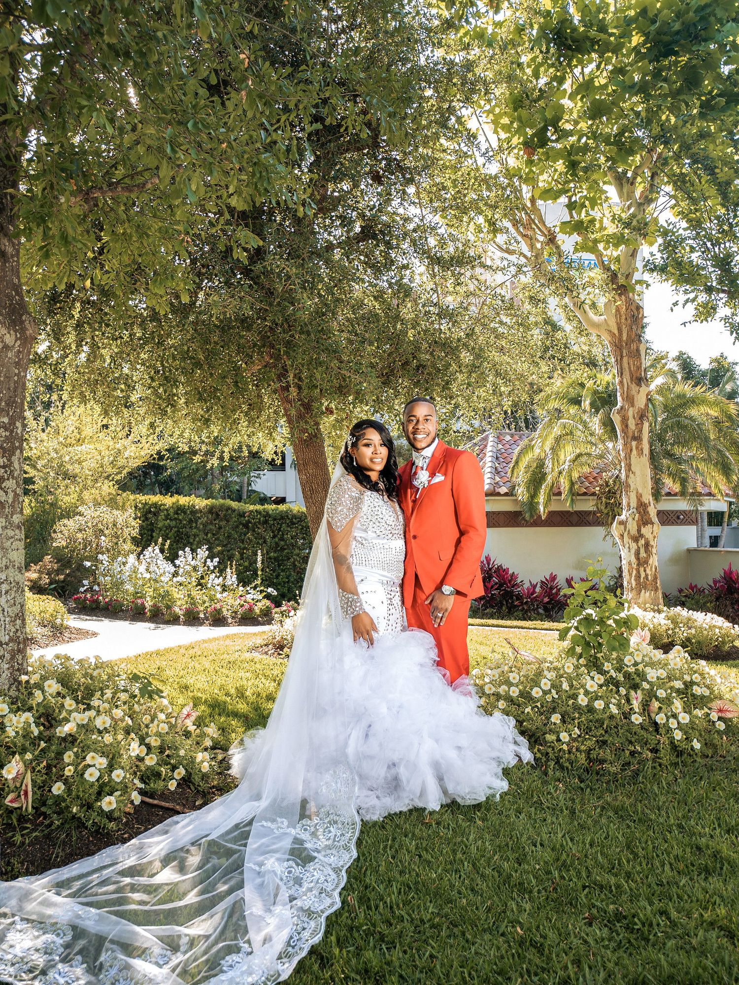 A wedding couple poses together in a lush garden setting with white floral decorations and a long trailing lace wedding gown.