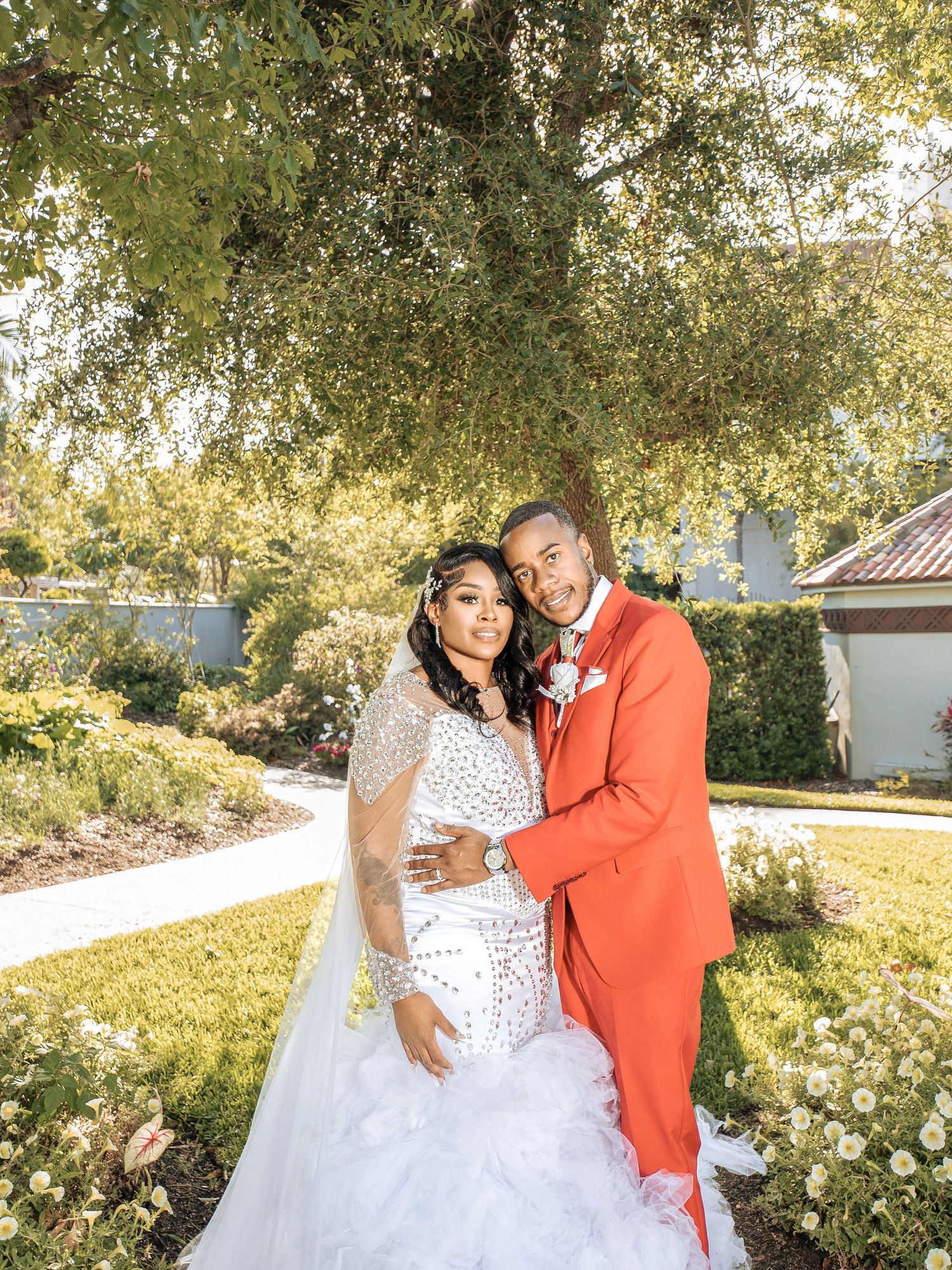 Wedding couple poses together outdoors with a dazzling white dress and bright orange suit under sunlit trees.