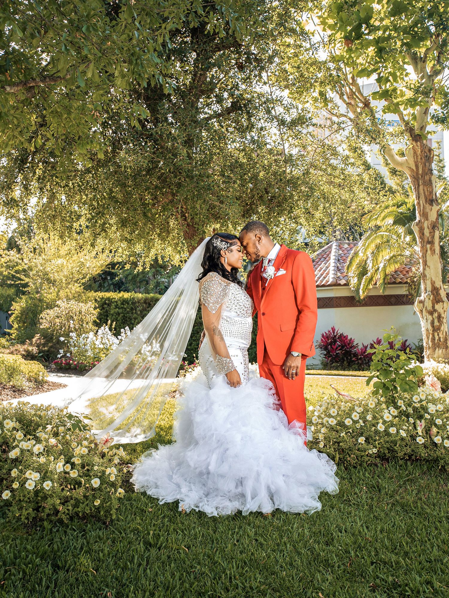 A bride in a white ruffled gown and groom in a bright red suit pose together under trees in a garden setting.