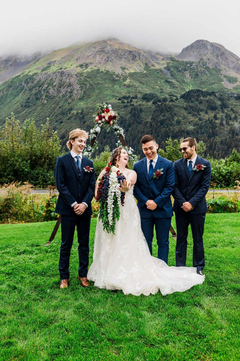 Small wedding group poses on grass with dramatic mountain peaks in background.