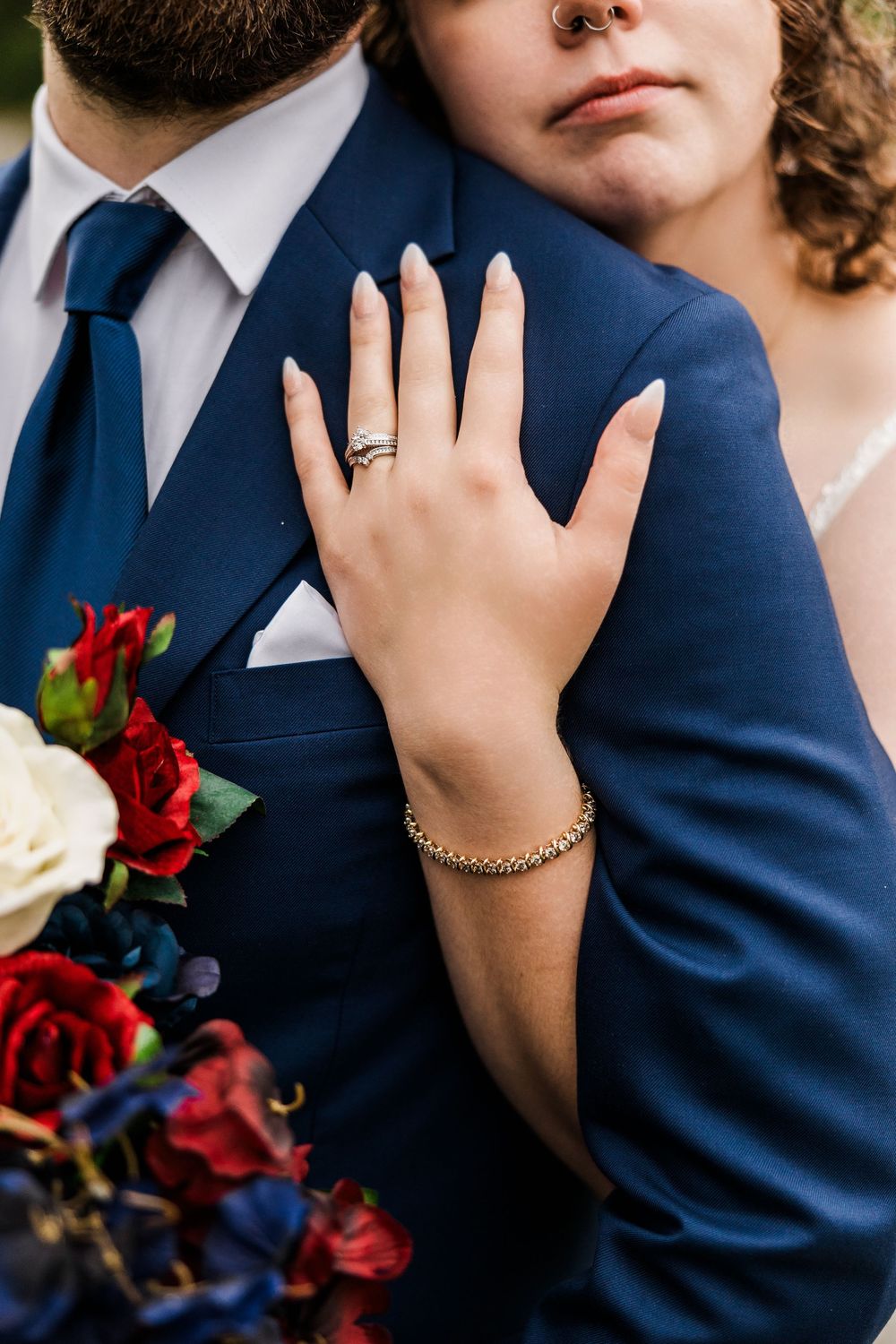 Wedding detail shot of hands with rings against navy suit and red bouquet flowers.
