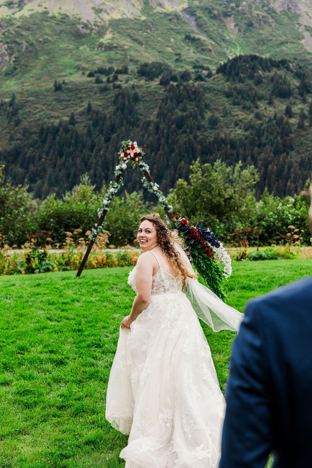Bride in white dress twirls in mountain meadow with floral arch and stunning landscape.