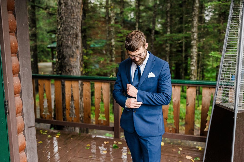 A person in a navy blue suit stands on a wooden deck surrounded by forest.