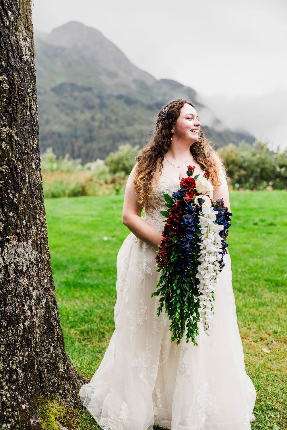 Bride holding cascading bouquet of red and blue flowers against mountain backdrop.