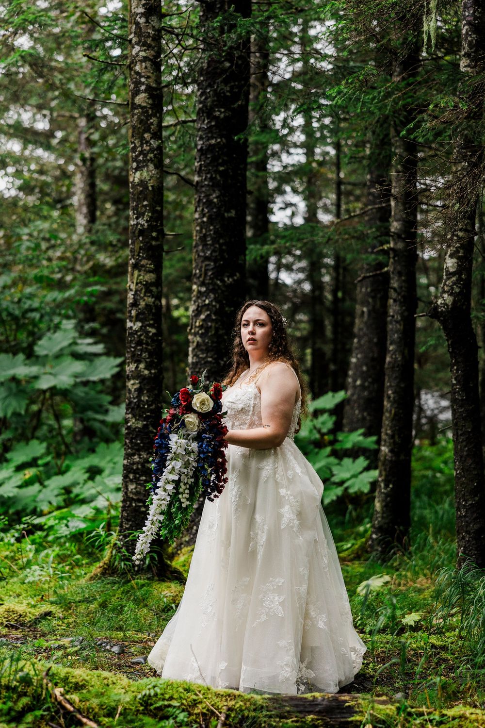 Bride in lace gown poses with bouquet in enchanted forest setting.