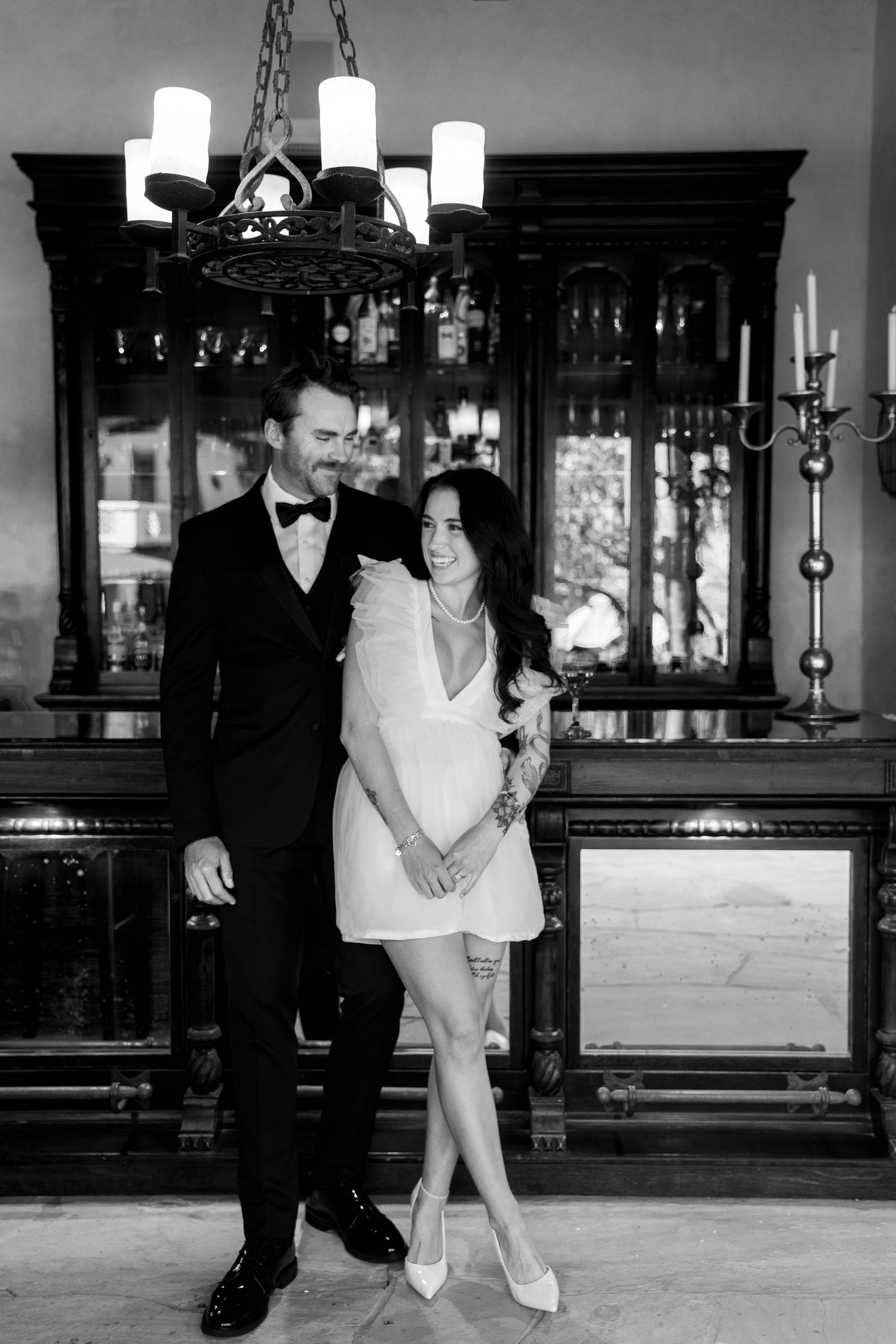 Elegant black and white photo of couple in formal attire posing in front of vintage wooden cabinets with chandelier.